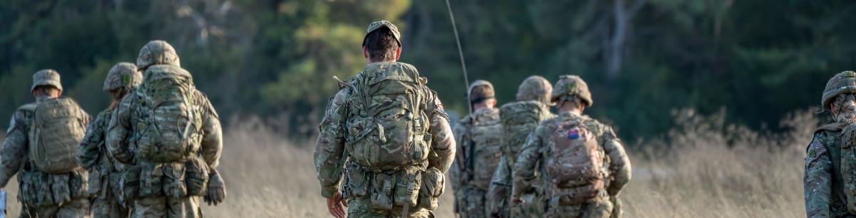 Armed Forces member in uniform walking through a field towards woods, shown from behind.