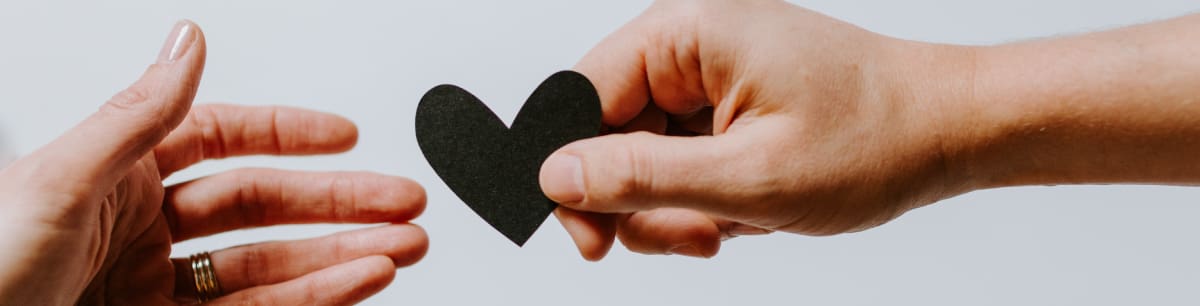 Two hands holding a black paper heart against a white background, only the hands are visible.