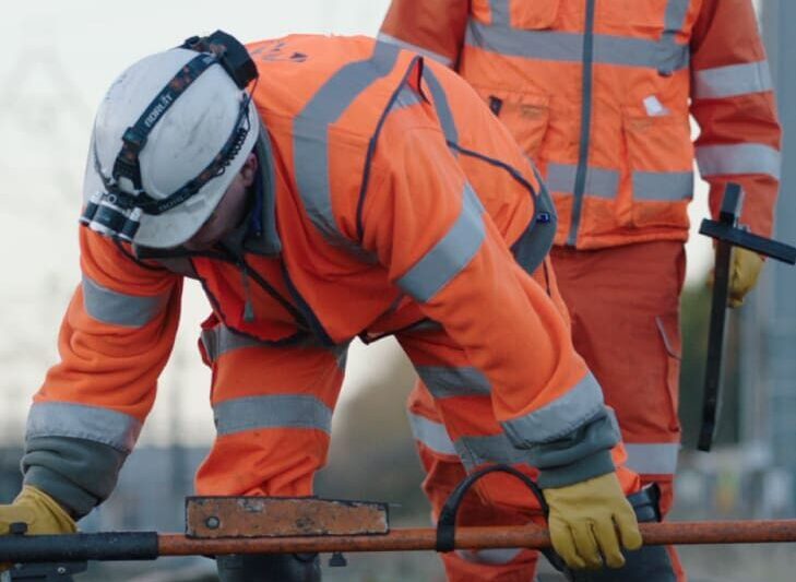 Two NRL rail workers inspect tracks, wearing full orange PPE and hard hats.