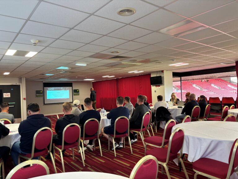 Group seated at tables listens to a presenter beside a screen in a conference room with red chairs.