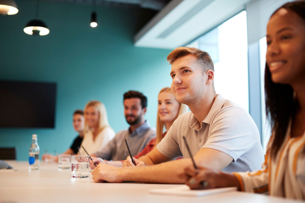 Five people sit at a table with notebooks and glasses of water, listening during a meeting in a bright room.