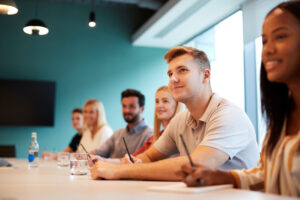 Five people sit at a table with notebooks and glasses of water, listening during a meeting in a bright room.