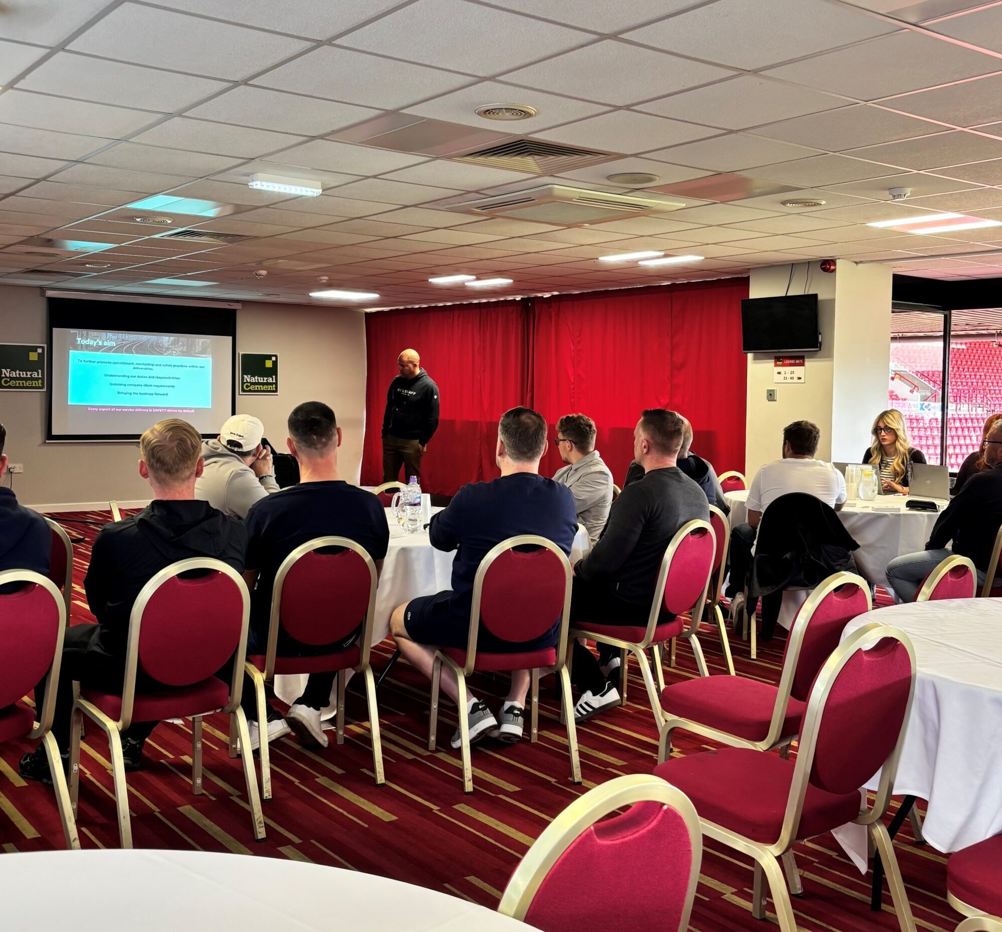 Group seated in a stadium lounge watching a safety presentation on a screen.
