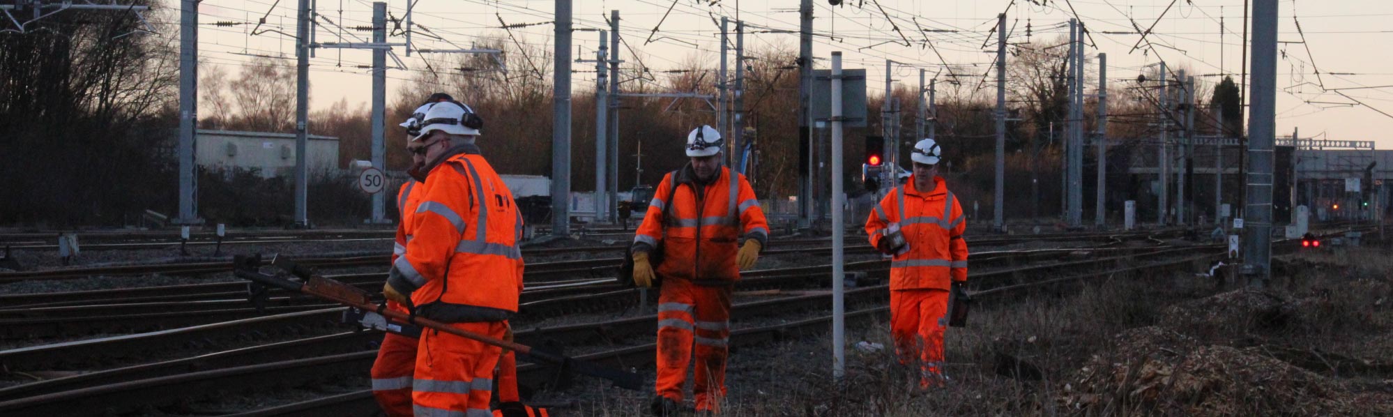 Rail workers in high‑visibility clothing walking beside tracks and overhead power lines during inspection work.