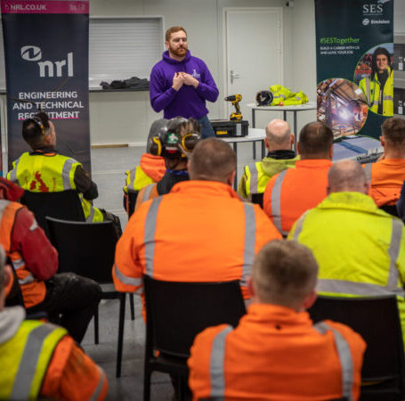 Person presents to a seated group in high-visibility clothing during a workplace safety briefing.
