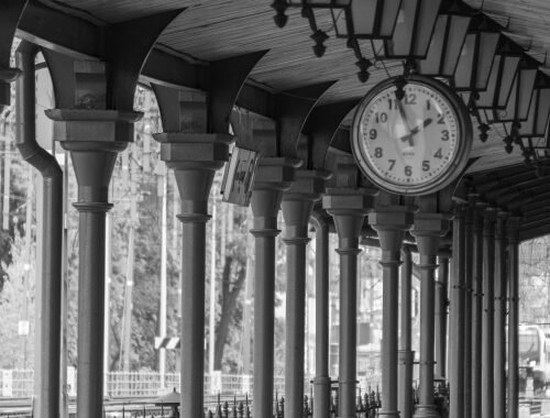 Clock mounted above a covered platform walkway with columns, looking along a railway station concourse.