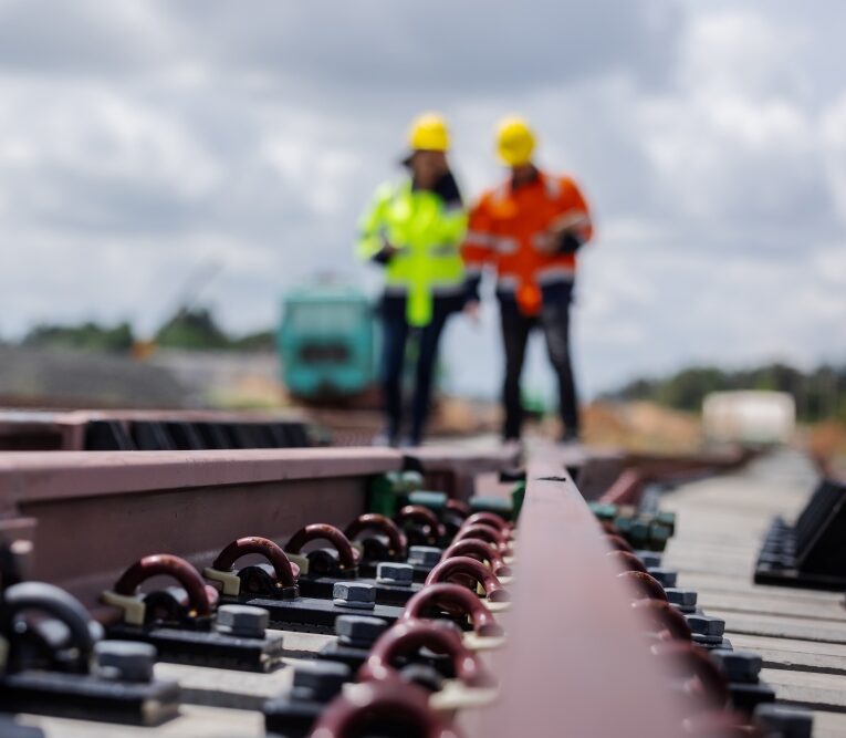 Close-up of railway track components with two workers in high-visibility clothing standing in the background.