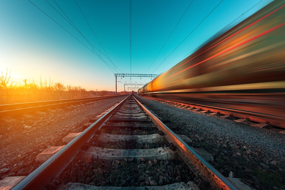 Blurred train speeds past on railway tracks with motion lines against a bright sunset sky.