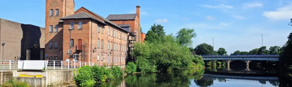 Historic red brick mill buildings beside a calm canal with trees and a bridge in the background.