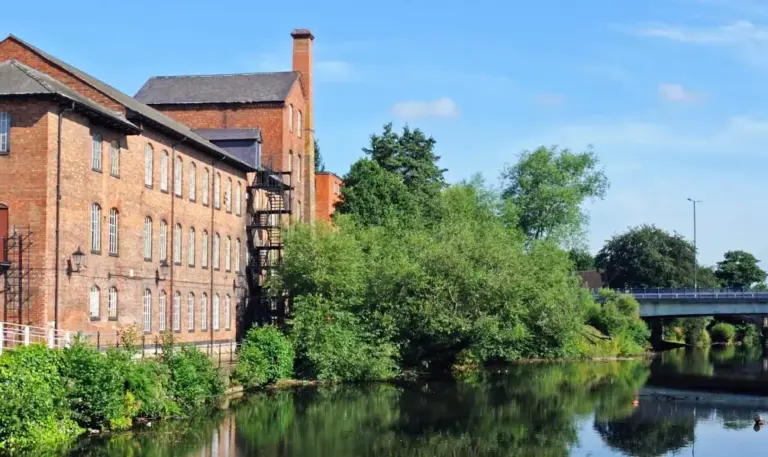Historic red brick mill buildings beside a calm canal with trees and a bridge in the background.