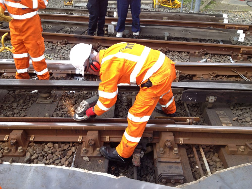 Worker in high-visibility clothing uses a grinder on railway track, producing sparks during maintenance.