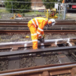 Worker in high-visibility clothing bends over rail using a handheld tool, with cables and equipment nearby.