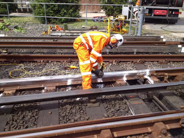 Worker in high-visibility clothing bends over rail using a handheld tool, with cables and equipment nearby.