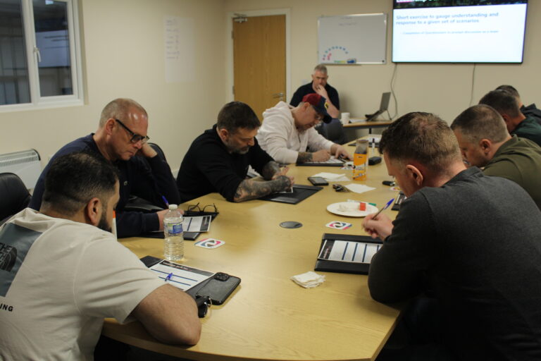 Group seated around a table writing notes while a presenter speaks at front of room.