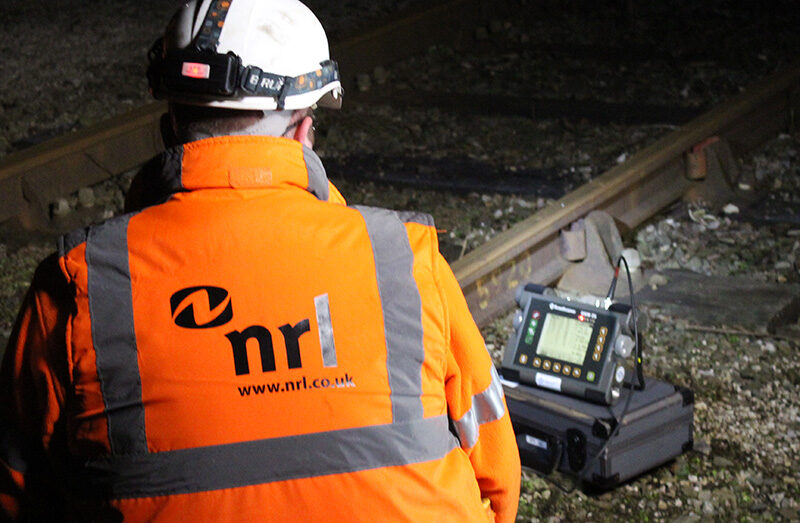 Worker in high-visibility clothing uses diagnostic equipment beside railway track in low light.