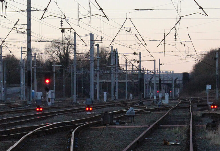 Multiple railway tracks with red signal lights and overhead cables leading towards a station.
