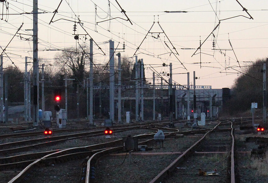 Multiple railway tracks with red signal lights and overhead cables leading towards a station.