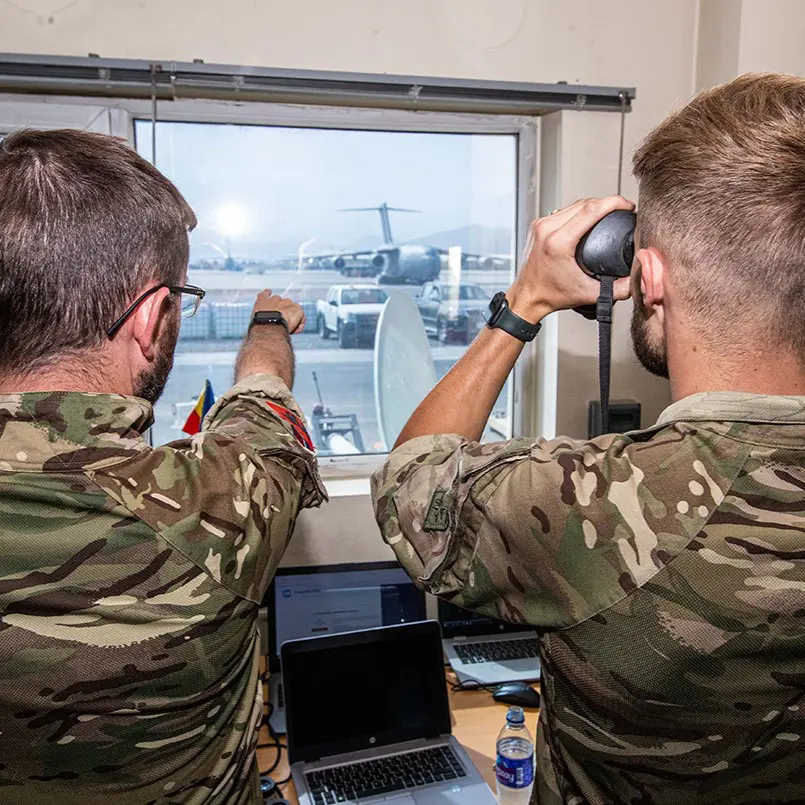 Two people in military uniform look out of a window, one using binoculars to view aircraft on a runway.