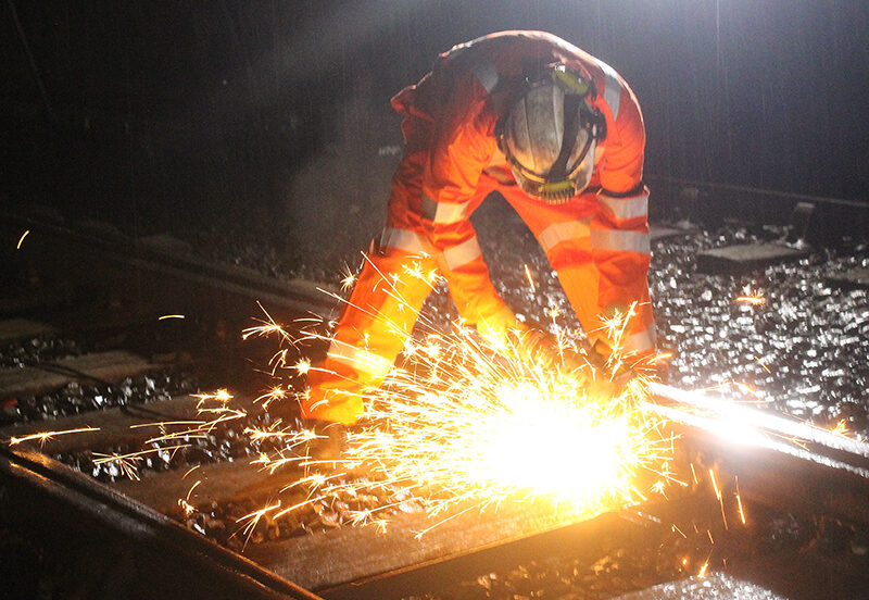 Worker in high-visibility clothing uses grinding equipment on rail, producing bright sparks at night.