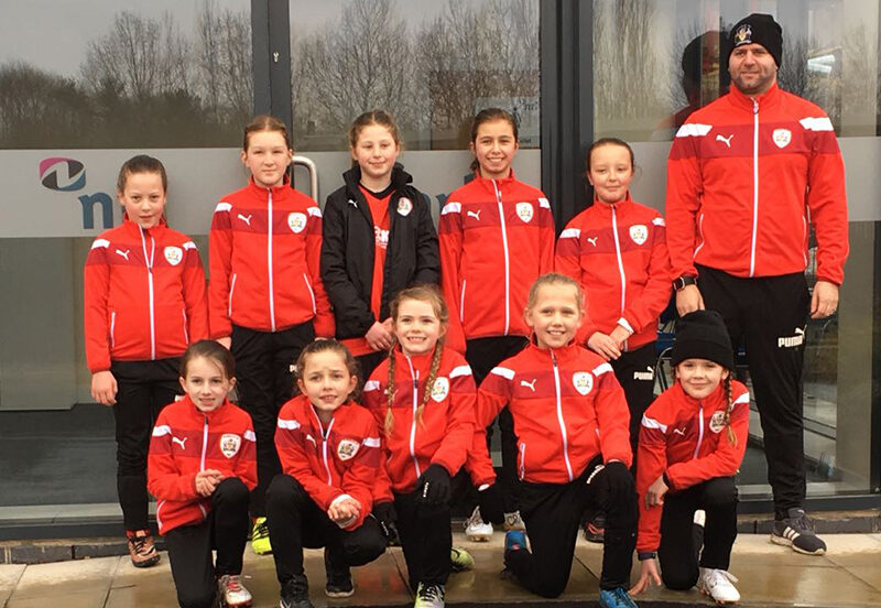 Group of children in matching red sports jackets pose with a coach outside a building.