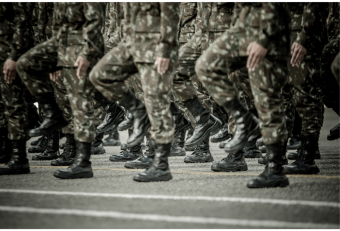 Rows of military personnel in uniform march in step, showing boots and legs in a parade formation.
