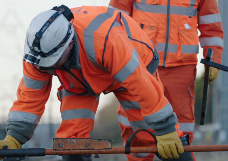 Worker in high-visibility clothing leans over railway track using a tool, with another standing nearby.