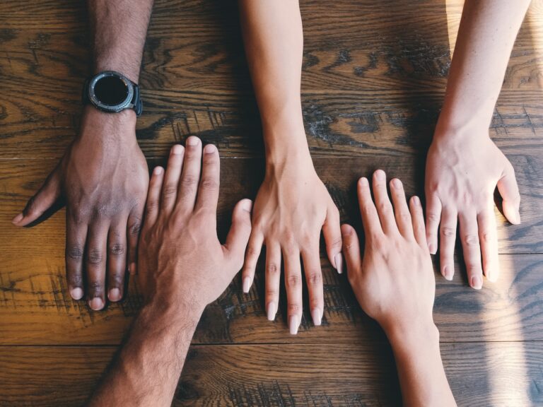Four hands placed flat on a wooden table, arranged side by side in a horizontal line.