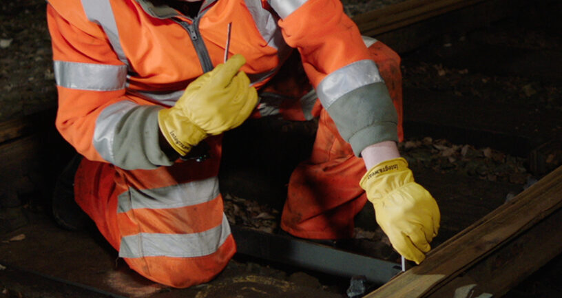 Worker in high-visibility clothing kneels beside a rail using a hand tool on track fittings.