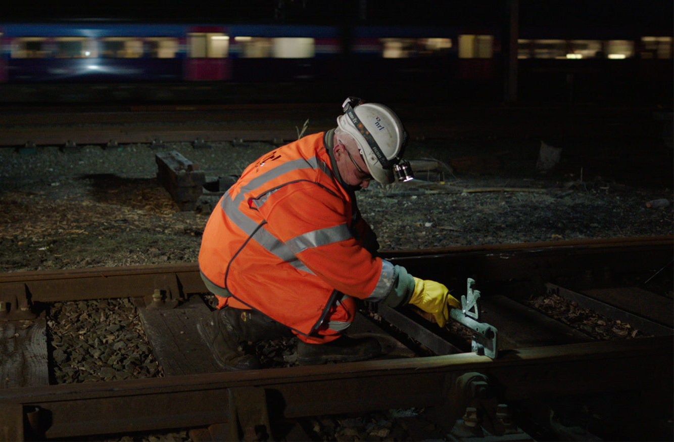 Worker in high-visibility clothing kneels on railway track at night, using a measuring tool.