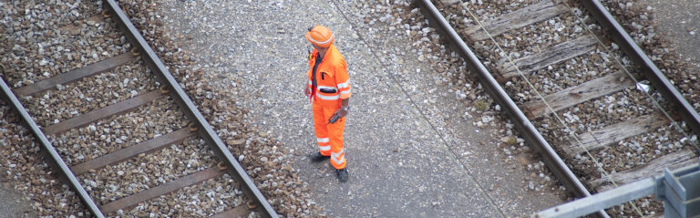 Worker in high-visibility clothing walks between parallel railway tracks on gravel ballast.