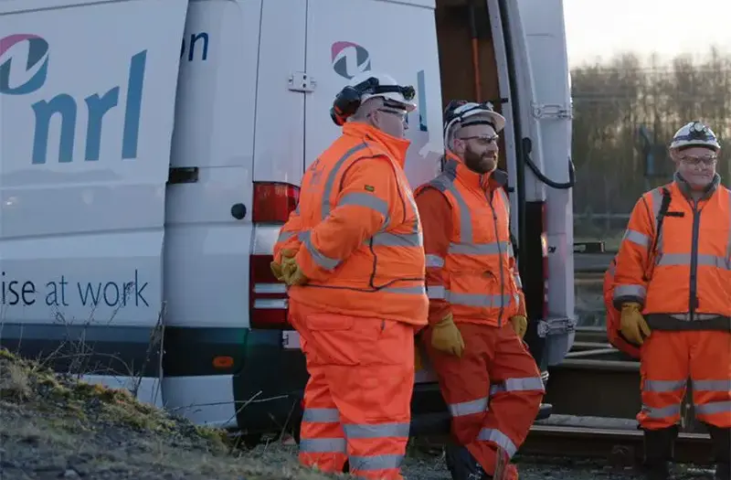 Three workers in high-visibility clothing stand beside an NRL van near railway tracks.