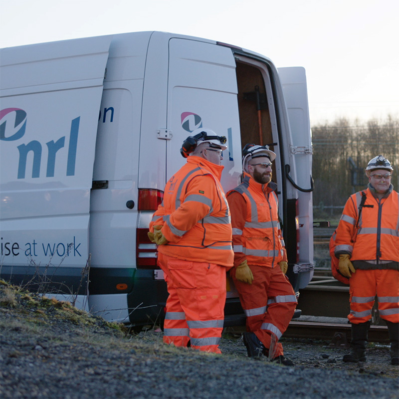 Three workers in high-visibility clothing stand beside an NRL van near railway tracks outdoors.
