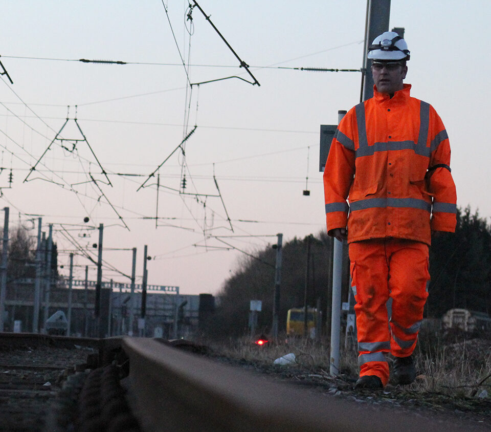 Worker in high-visibility clothing walks beside railway track with overhead lines and signals.