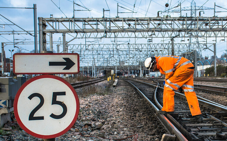 Worker in high-visibility clothing inspects railway track beside a 25 speed limit sign and signal gantry.