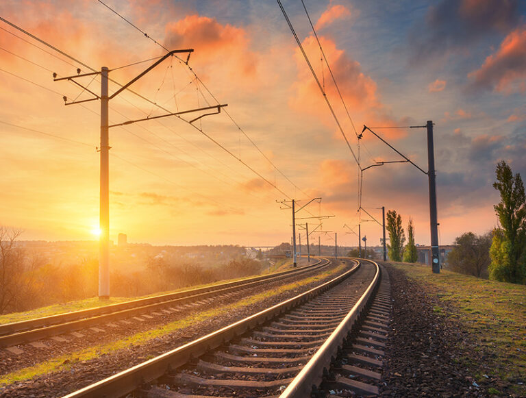 Railway tracks curve into the distance under overhead lines with a colourful sunset sky.