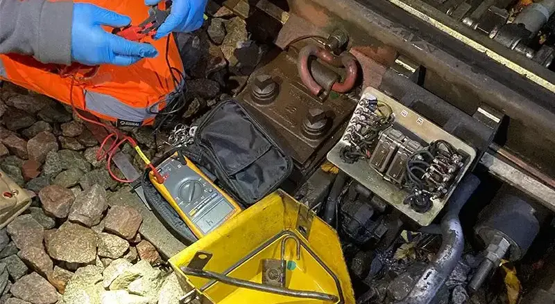 Worker in high-visibility clothing uses testing equipment connected to railway track components.