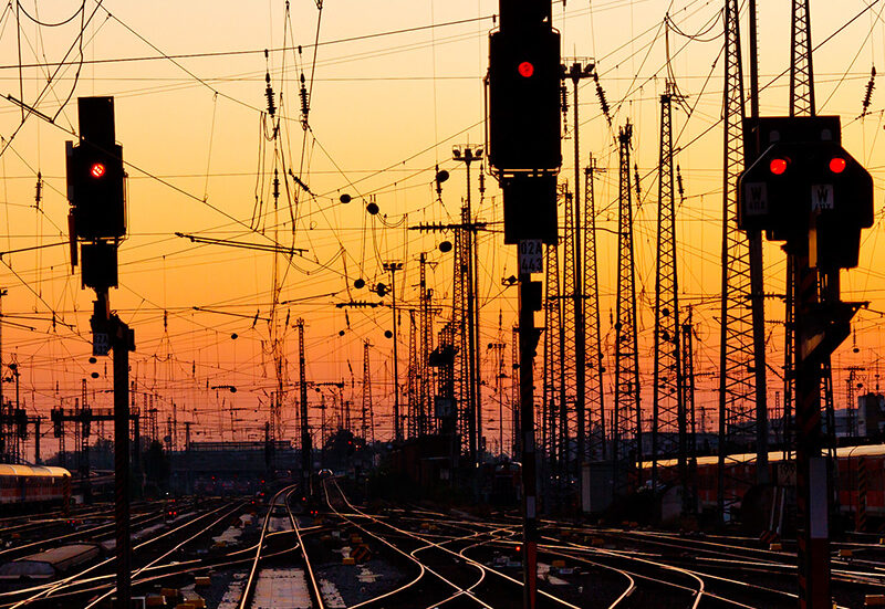 Railway tracks with signal lights and dense overhead cables silhouetted against an orange sunset sky.