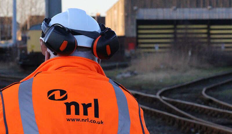 Person in high-visibility clothing with NRL logo stands beside railway tracks wearing safety helmet and ear protection.