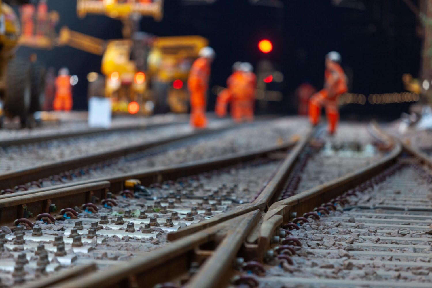Railway tracks in foreground with workers and machinery blurred under lights during night work.