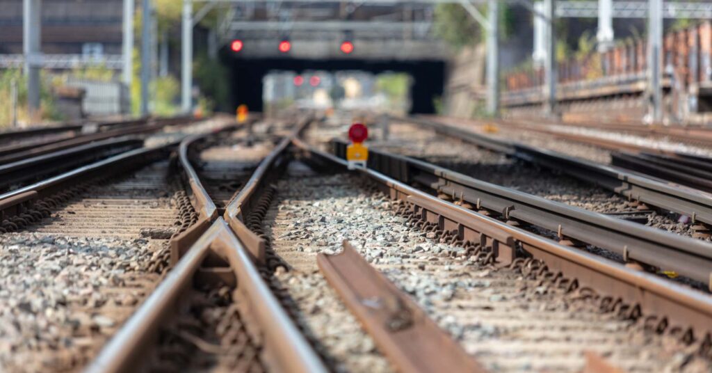 Multiple railway tracks split and merge with a signal, leading toward a bridge in the background.