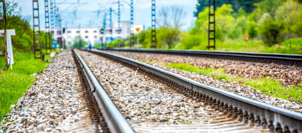 Two parallel railway tracks on gravel ballast with overhead lines and greenery along the side.
