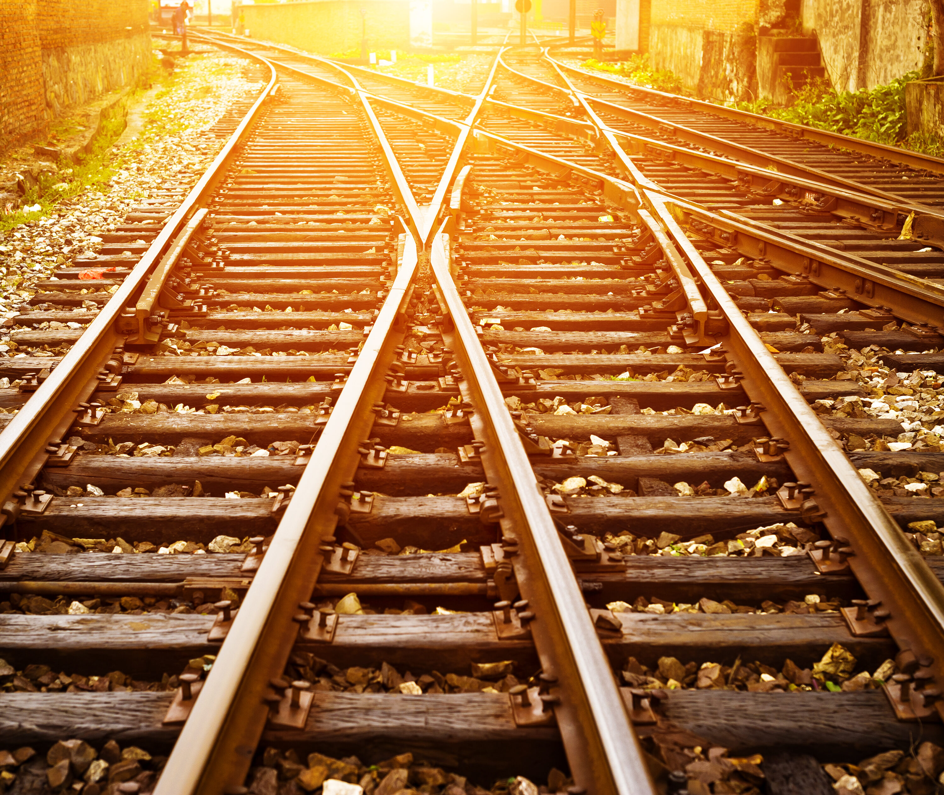 Multiple railway tracks split and cross at a junction, lit by warm sunlight between brick walls.