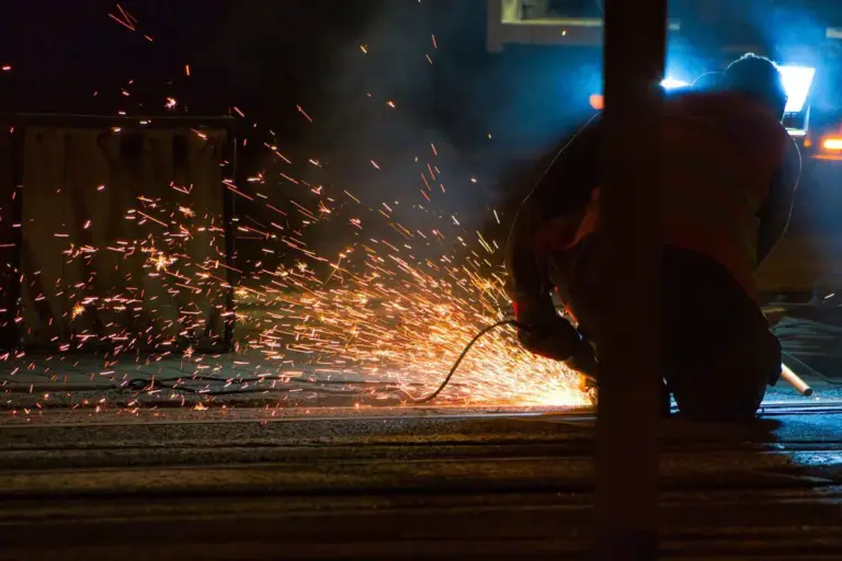 Worker operates rail grinder producing bright sparks on track during night maintenance.
