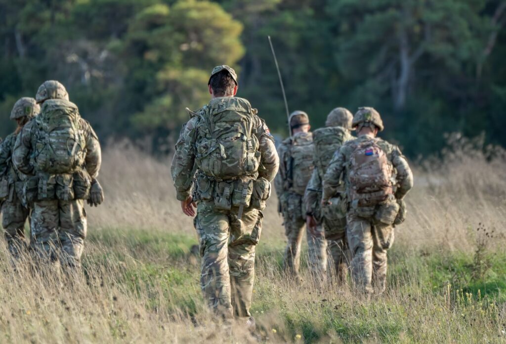 Group of military personnel in uniform walk through tall grass carrying backpacks in a rural area.