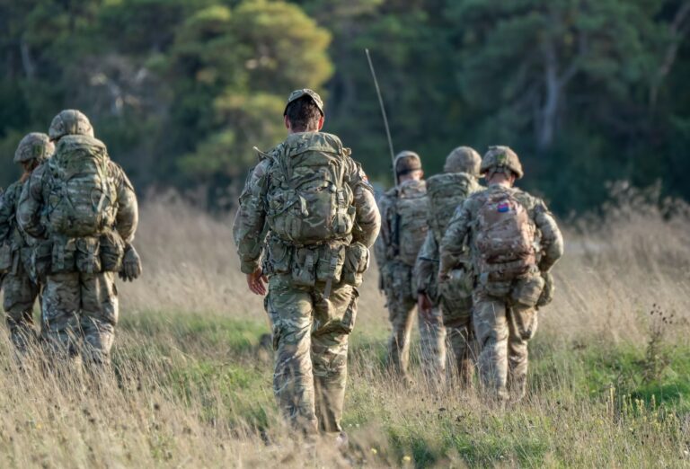 Group of military personnel in uniform walk through tall grass carrying backpacks in a rural area.